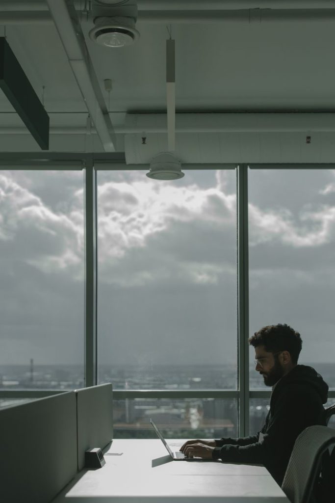 Man in a black jacket sitting at a desk and working on a laptop in a bright modern urban office with glass windows.